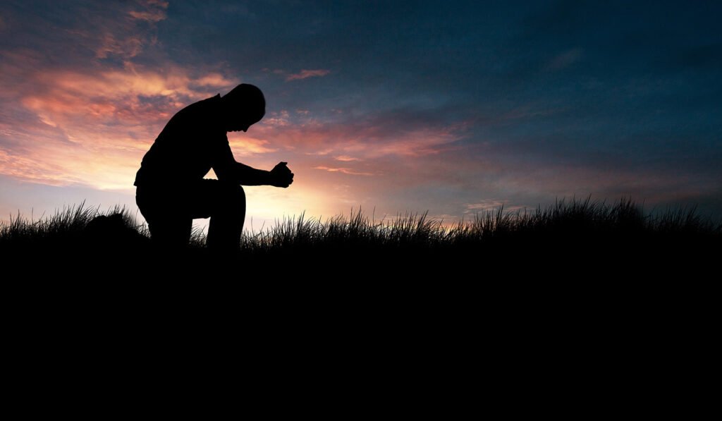 Man praying in a grassy field with a sunset behind him. Concept: reverence, prayer, love of God.